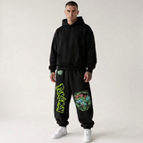 A man modeling a black pullover hoodie and matching black jogger sweatpants with colorful graphic prints, including a large green 'Pokémon' logo on one leg and a multicolor turtle graphic on the other, against a plain light gray studio background.