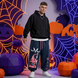 A man stands in a Halloween-themed studio setting wearing dark navy sweatpants with a graphic of a pink demon character and Japanese text, paired with a black hoodie and white sneakers.