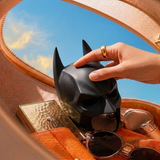 A black Batman-style mask is being held by a hand inside an open tan leather bag, surrounded by sunglasses, a gold compact, and a blue sky visible through the bag's opening.