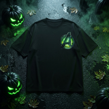 A black short-sleeve t-shirt featuring a glowing green and black Avengers 'A' logo on the left chest, displayed in a Halloween-themed flat-lay setting with jack-o'-lanterns and wet leaves.