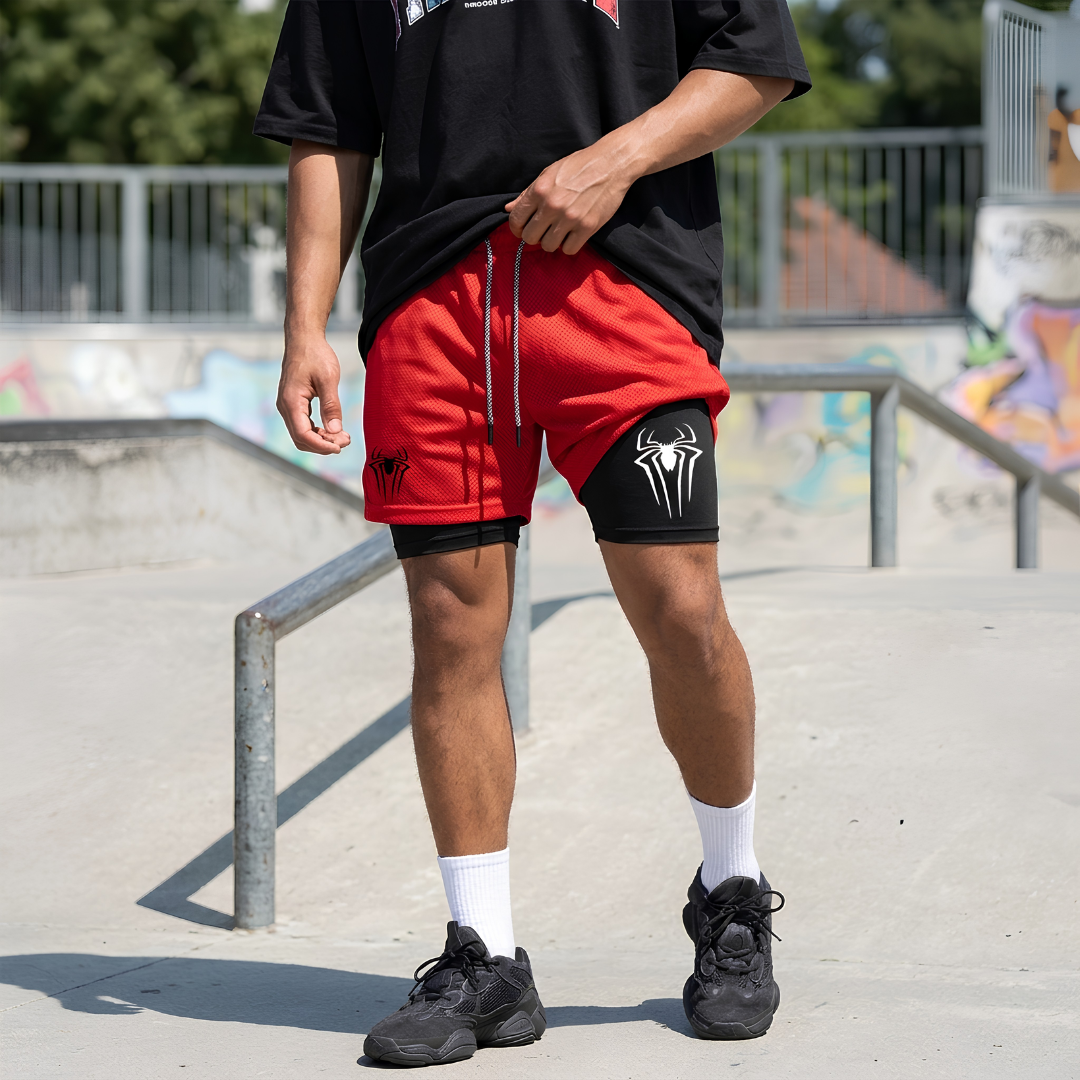 A person standing at a concrete skatepark wearing red athletic shorts with black spider-themed graphics, a black t-shirt, white socks, and black sneakers.