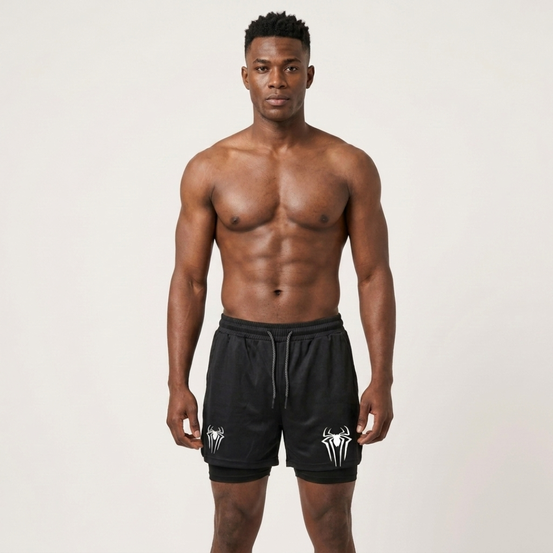 A shirtless man modeling black athletic shorts with white spider logos on the left leg, standing against a plain light gray studio background.