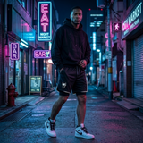 A man stands on a city street at night wearing a black hoodie and black shorts with white sneakers. Neon signs for bars and a market glow in blue, pink, and purple tones around him.