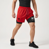 A man modeling red and black dual-layer athletic shorts with Spider-Man themed logos, paired with a black shirt and black sneakers, against a light gray studio background.
