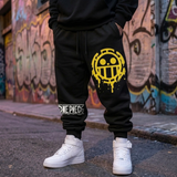 A person wearing black One Piece jogger sweatpants with a large yellow anime logo on the thigh and a white logo band near the cuff, paired with white sneakers, standing on a street in front of a graffiti-covered wall.