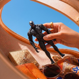 A hand holds a black Venom action figure above an open tan leather purse containing sunglasses, a gold compact, and a patterned scarf, with blue sky visible in the background.
