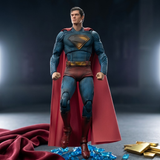 Superman action figure or collectible statue in a classic blue and red suit with a flowing red cape, standing on a dark surface with blue glass pieces and gold bars around it.