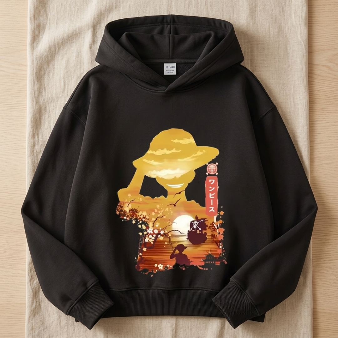A black pullover hoodie laid flat on a beige fabric surface, featuring a large front graphic of a golden hat silhouette with a sunset scene, cherry blossoms, and vertical Japanese text.
